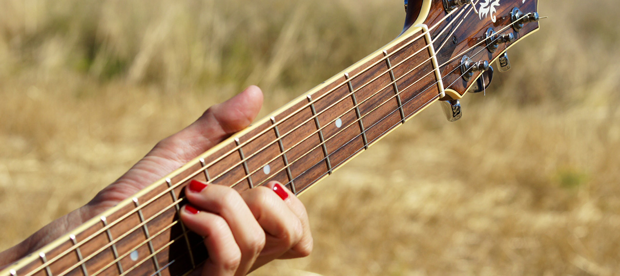 Mano con las uñas pintadas de rojo tocando el mástil de una guitarra acústica en un campo de trigo