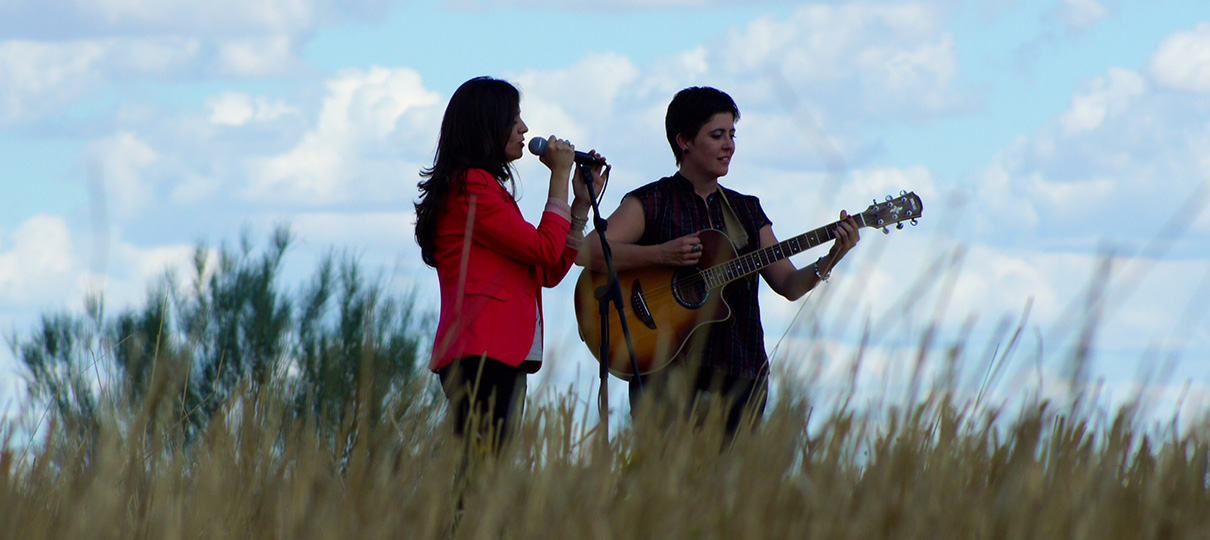 Una de ellas canta al micrófono y la otra toca la guitarra acústica en medio de un campo de trigo bajo un cielo con nubes